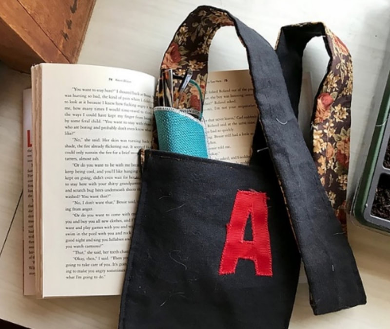a black fabric shoulder bag decorated with a red letter A, displayed with an open copy of the Scarlet Letter