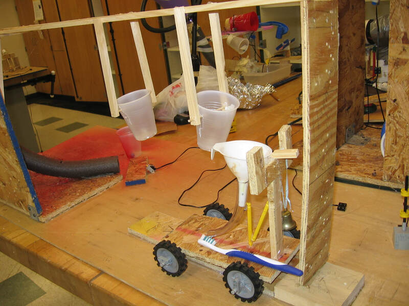 portion of a Rube Goldberg machine, showing a vehicle triggered by something that drops through a funnel