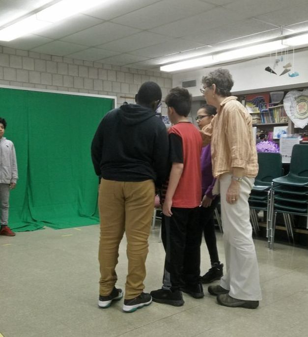 3 students and a teacher cluster around a camera, shooting a 4th student against a green screen