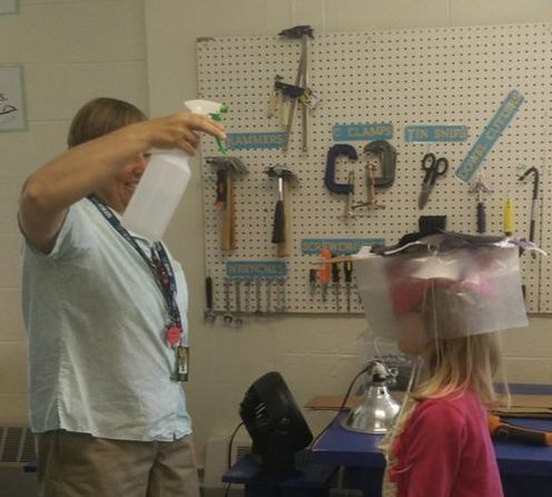 a student wears a weather-proof hat while a teacher sprays  water on her to test it 