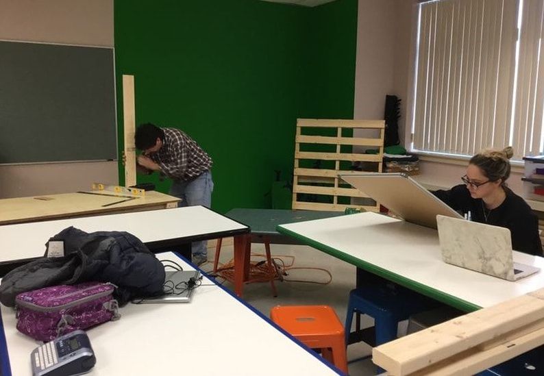 Photo of two students working on large tables, one with wood an another with a laptop